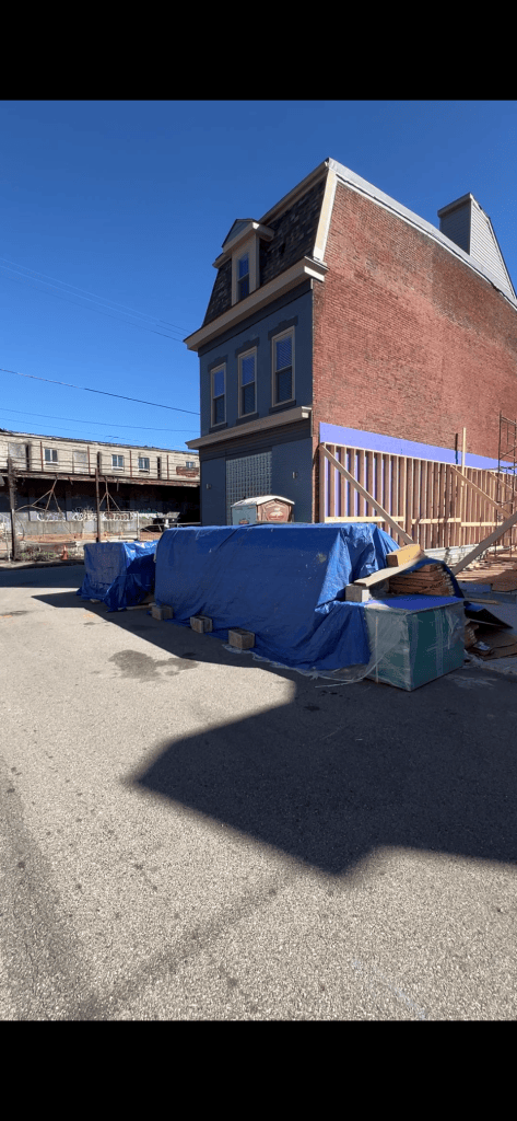 A side view of a brick building under renovation with blue tarps covering construction materials and debris in front of it, set against a clear blue sky.