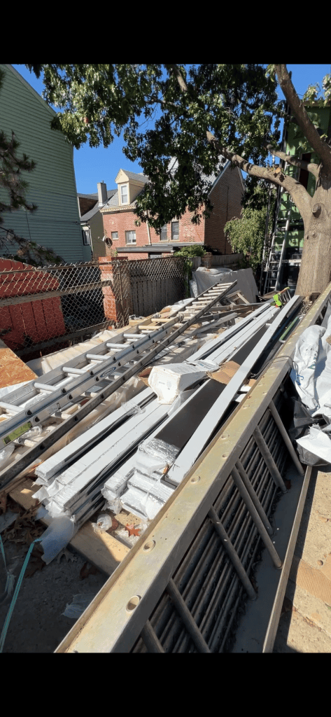 A construction site with a pile of metal beams, ladders, and various construction materials, surrounded by trees and fences.