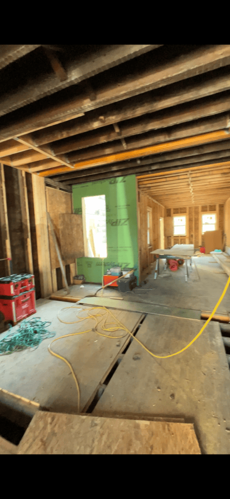 Interior of a partially constructed house showing wooden beams, tools, and construction materials, including a green wall section and a table.